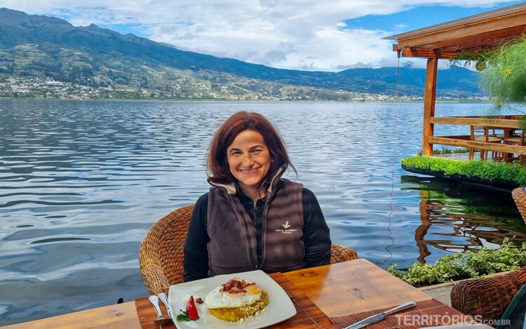 Mulher morena de cabelos curtos sorri sentada em mesa na frente de lago. Um prato com ovos e bacon é servido. Ela veste colete marrom e blusa preta.