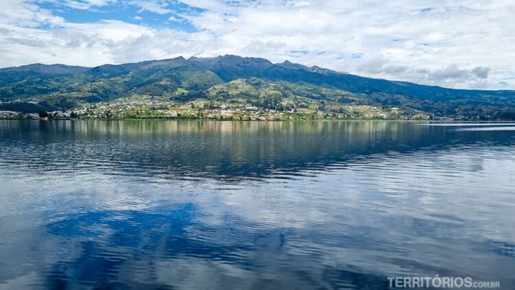 Lago San Pablo com reflexo do céu e a outra margem com montanhas e casas na zona rural de Otavalo