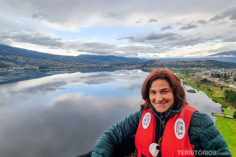 Mulher morena com salva-vidas e jaqueta verde sorri do alto do balão. Ao fundo, lago, montanhas e gramado.