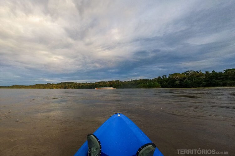 Pés sobre caiaque azul navega no rio Napo em direção a um barco ao longe. Céu com nuvens, mata verde nas margens do rio de cor marrom