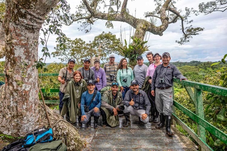 Grupo de turistas e guias faz pose para fotos encostados em cerca do mirante. Árvore de tronco grosso a frente, céu com nuvens abundante vegetação verde