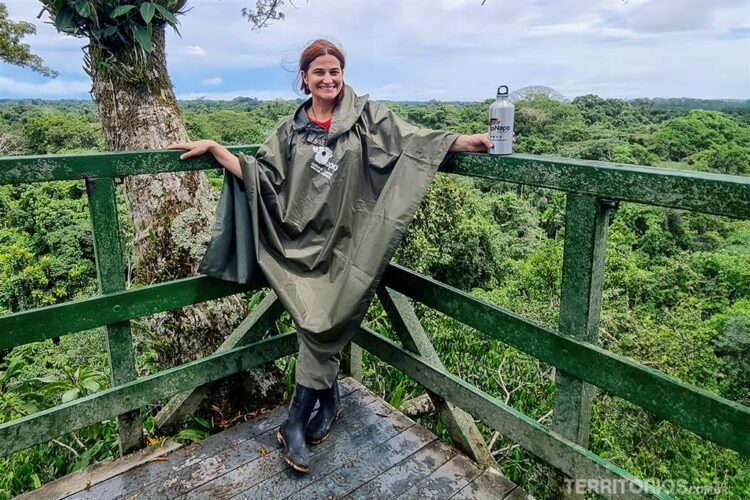 Mulher sorri e faz pose encostada em cerca do mirante. Veste poncho verde, botas de borracha e segura garrafa metálica na mão esquerda