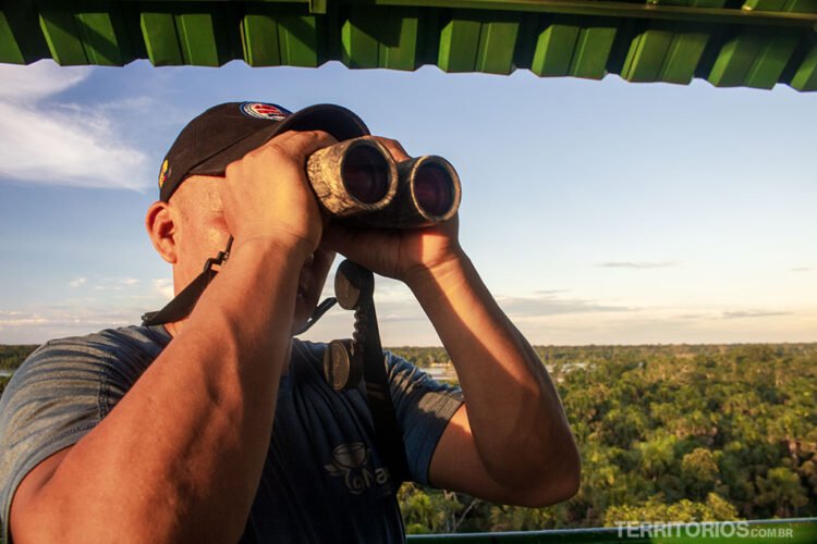 Indígena de Napo olha pelo binóculo em mirante para a selva. Ele tem pele bronzeada, veste blusa cinza e boné preto