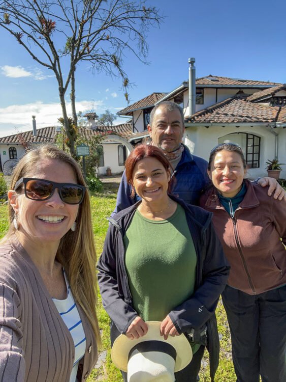 Três mulheres e um homem fazem pose em frente a casa branca em dia ensolarado com céu azul e poucas nuvens.