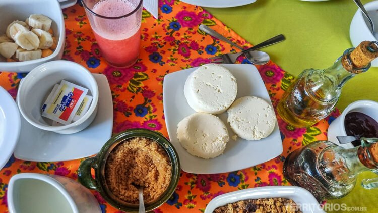 Mesa posta no café da manhã da Sierra Alisos com queijos, azeite, suco de melancia, açúcar, banana e picada. Cerâmicas brancas em toalha florida em tons rosa, laranja e azul. Toalha verde ao fundo.