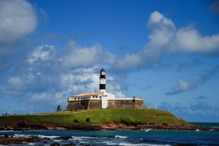 Farol da Barra sobre ilha com gramado verde rodeada pelo mar e céu azul