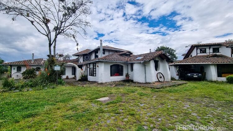 Sierra Alisos: casa branca com gramado verde e telhado vermelho com jardim na frente. Céu azul entre nuvens.