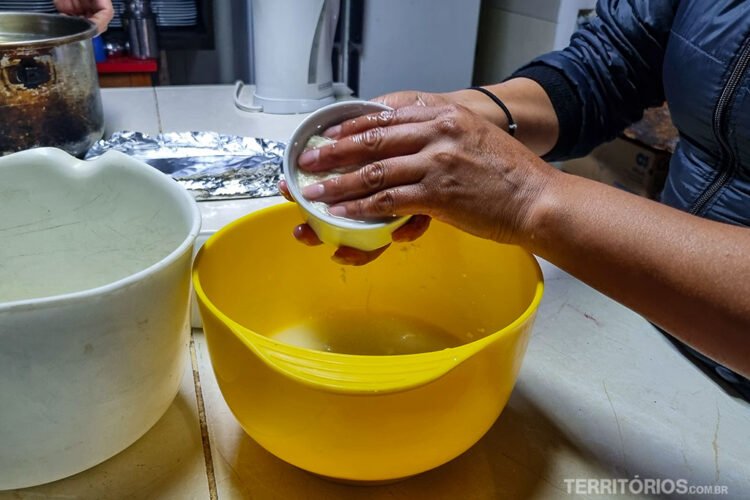 Mãos pressionam coalhada com líquido em pote de cerâmica branco, líquido cai em pote maior de cor amarela. 