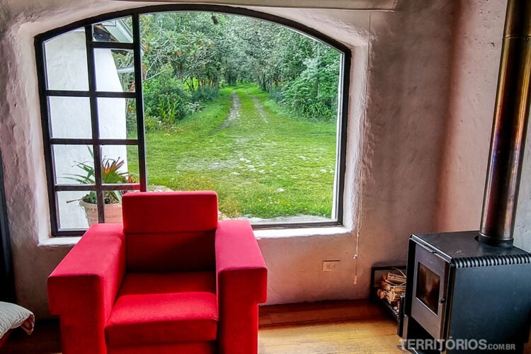 Poltrona vermelha em frente a janela com vista para o campo verde e jardim. Parede branca, mesa com lenha e aquecedor a lenha no Hotel de Campo Sierra Alisos