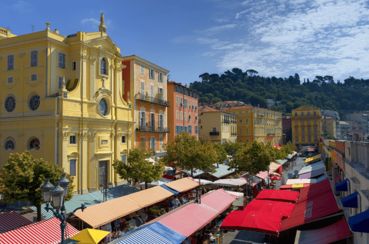 Mercado local com toldos coloridos em cidade com prédios antigos, céu azul com poucas nuvens.