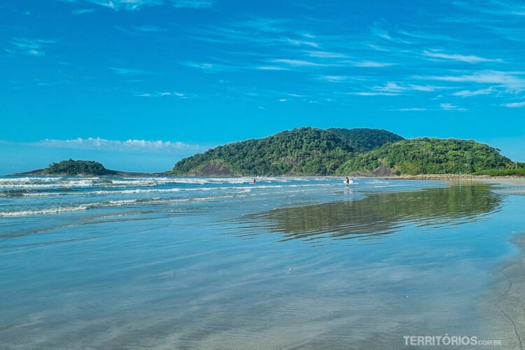 Lado sul da praia Riviera de São Lourenço. Surfistas entram no mar em dia azul com poucas nuvens. Montanhas verdes com rochas ao fundo