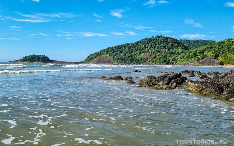 Água do mar da praia Riviera de São Lourenço na maré enchendo. Montanhas verdes e rochas ao fundo, céu azul com poucas nuvens