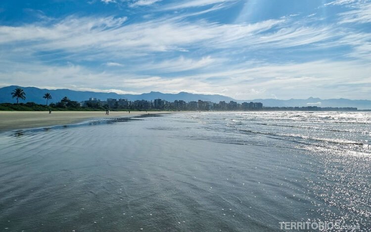 Vista para a Serra do Mar, coqueiros, areia e prédios da beira da praia Riviera de São Lourenço. Céu azul com muitas nuvens