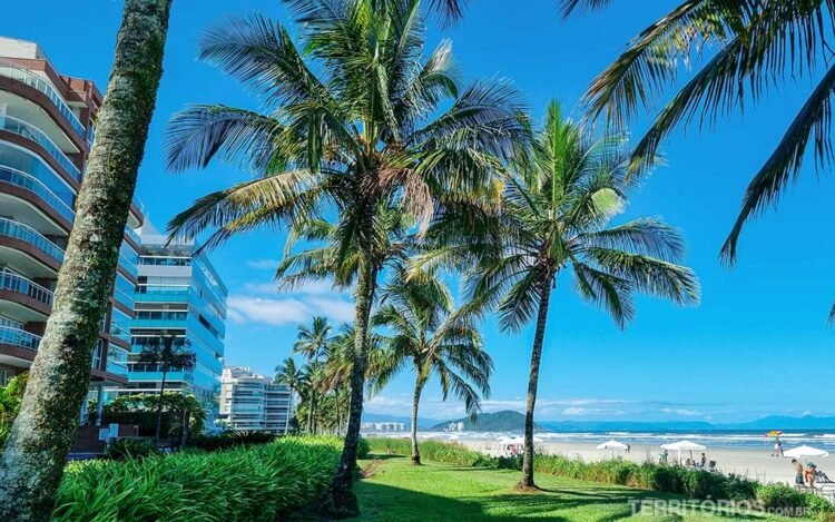 Prédios na beira-mar da Riviera de São Lourenço, jardim com coqueiros e gramado verde, faixa de areia e algumas barracas. Céu azul com poucas nuvens, montanhas distantes e pedaço do mar azul.