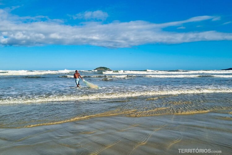 Pescador entra mo mar com tarrafa. Algumas ondas, céu azul, uma nuvem e ilha no centro da foto.