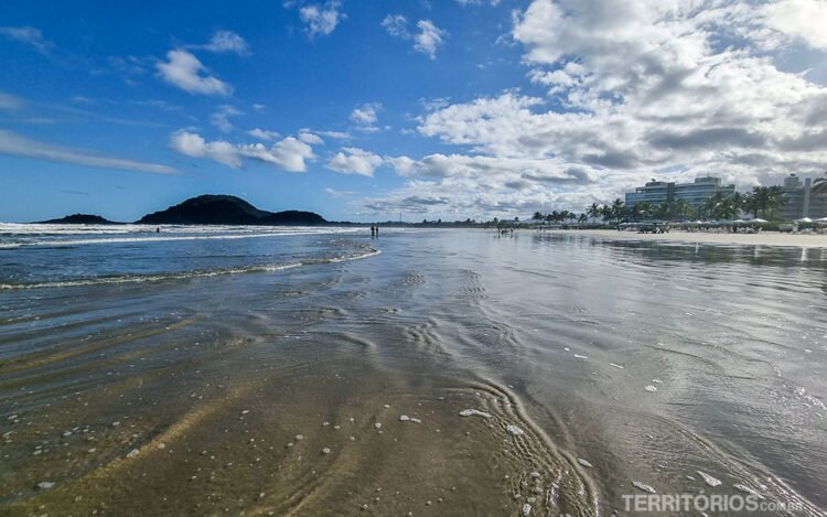 Praia Riviera de São Lourenço, vista da beira-mar para montanhas e prédios. Céu azul com nuvens.