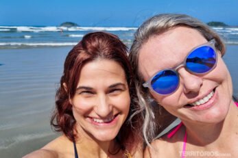 Duas mulheres, uma de óculos, sorriem com mar e ilhas ao fundo. Céu azul.