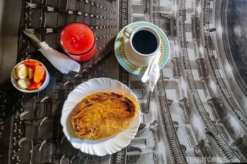 Mesa estampada com foto posta com xícara de café, prato com pão prensado, tigela de frutas, suco de melancia e talheres.