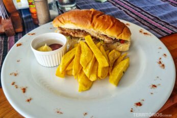 Sanduíche com batatas assadas servido em prato branco com temperos decorando e pote branco com molhos.