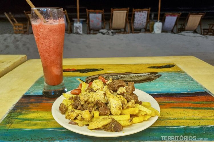 É noite na praia com cadeiras de costas. Suco natural em copo, carnes, ovos e batatas-fritas servidas em prato branco e mesa colorida de um bar em Isabela.