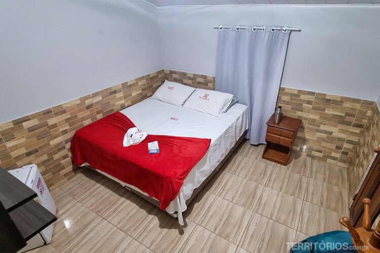 Suíte de hotel com paredes, roupa de cama e cortinas brancas, azulejos em tons de beje, bidê de madeira