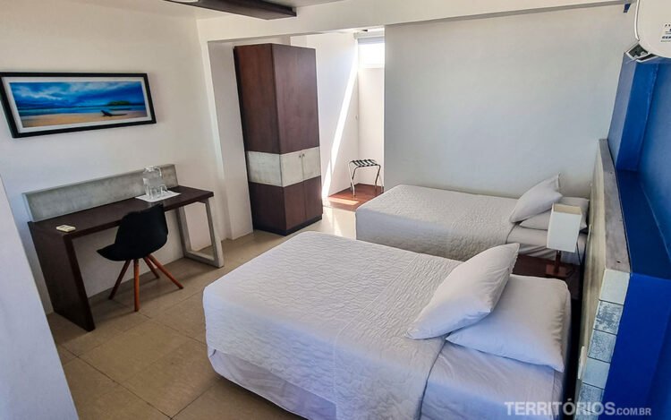 Suíte de hotel com duas camas de solteiro e roupa de cama branca. Móveis de madeira e quadro azul na parede branca.