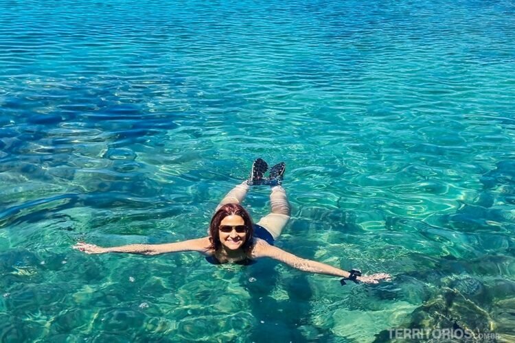 Mulher branca de óculos e cabelos escuros mergulha em mar cristalino em tons de verde.