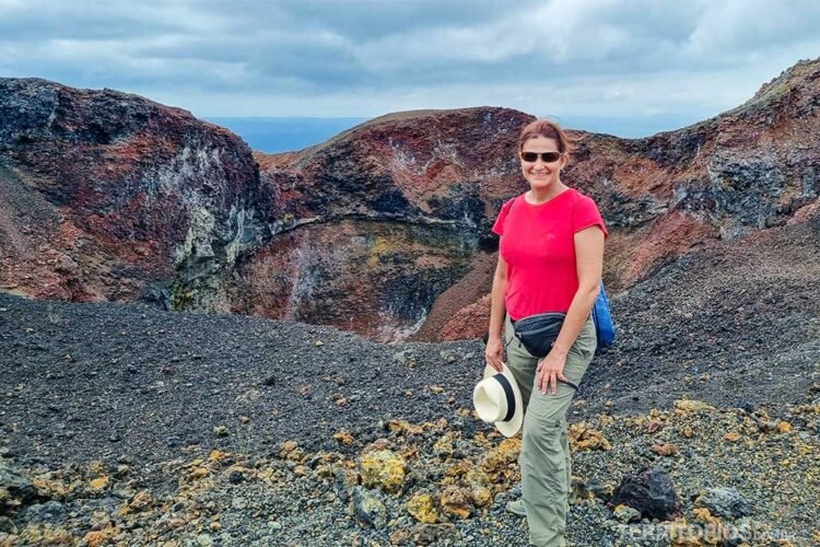 Mulher morena de óculos escuros veste blusa rosa, calça bege, pochete preta e leva chapéu de palha na mão. Esta em frente a uma cratera de vulcão em Isabela. Céu entre nuvens.