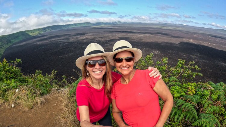 Duas mulheres, uma loira e outra morena, vestem camiseta rosa, óculos de sol e chapeu de palha. 
Estão em frente a uma cratera de vulcão na ilha Isabela