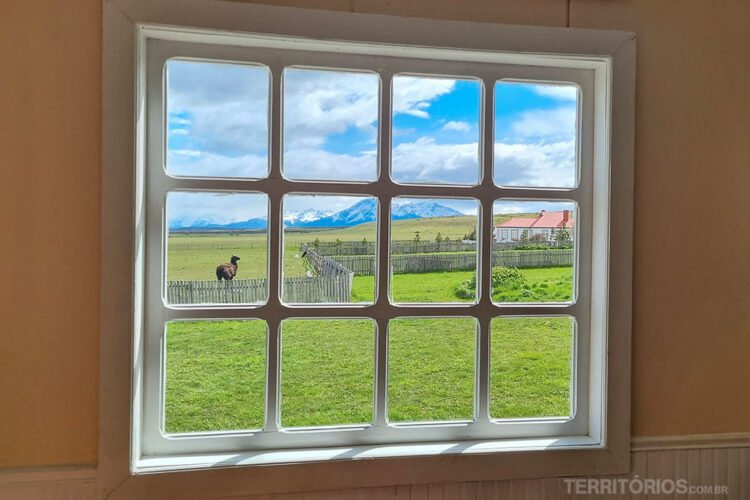 Janela em parede amarela tem vista para campo verde com cerca, uma olha, uma casa ao longe e montanhas nevadas. Céu azul com nuvens.