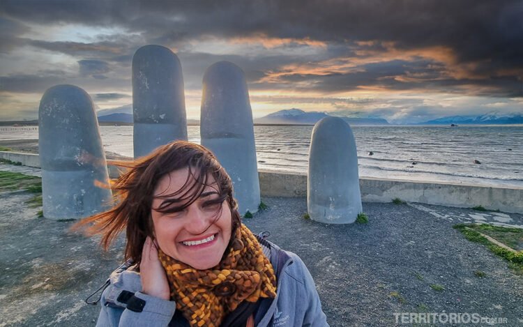 Mulher sorri segurando os cabelos ao vento em frente a escultura de mão gigante em orla de Puerto Natales. Céu azul com nuvens pesadas e luz no horário do pôr do sol.