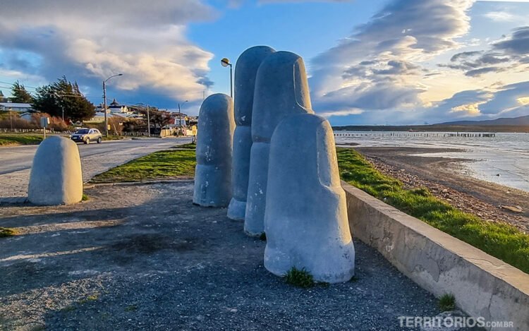 Escultura de mão gigante em orla de Puerto Natales. Céu azul com nuvens pesadas e luz no horário do pôr do sol