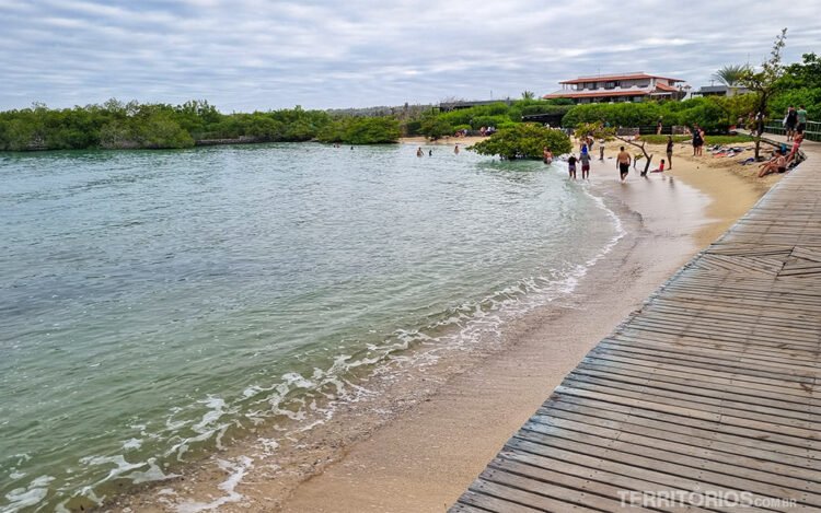 Passarela de madeira leva para praia com curta faixa de areia onde pessoas caminham em Santa Cruz. Mar em tons verde rodeado por vegetação verde e céu nublado. Um sobrado ao fundo direito.