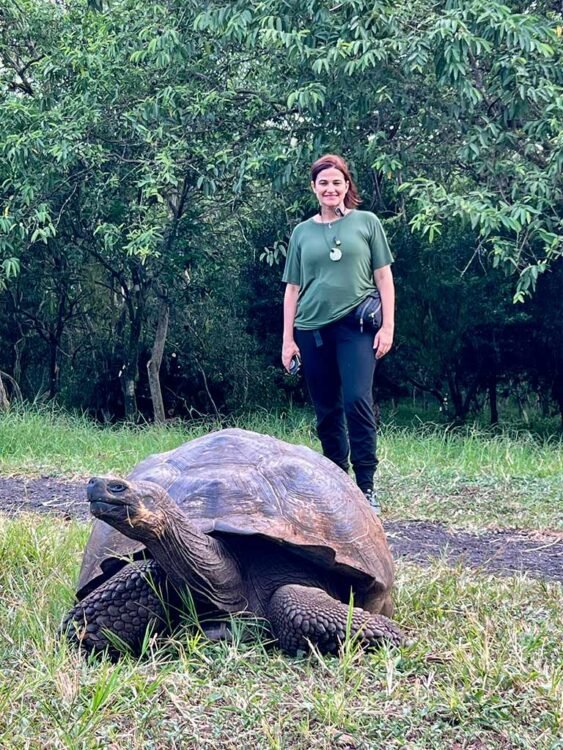 Tartaruga-gigante de Galápagos, na Ilha Santa Cruz, posicionada em frete a mulher morena de cabelos presos, blusa verde e calças pretas. Vegetação verde ao fundo.