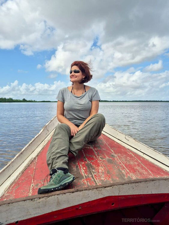 Mulher de cabelos escuros, óculos de sol, camiseta cinza, calças e tênis verde sentada na proa de uma barco a caminho da Ilha de Santana