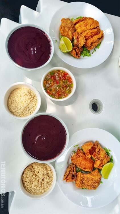 Pratos brancos servidos com peixe frito e limão em mesa branca de plástico. Potes brancos servidos com farinha, açaí e tempero na Ilha do Combú