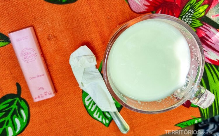 Xícara servida com leite em mesa com toalha laranja, um talher coberto com guardanapo e barra de chocolate embalada sobre mesa com toalha laranja estampada com verde e rosa