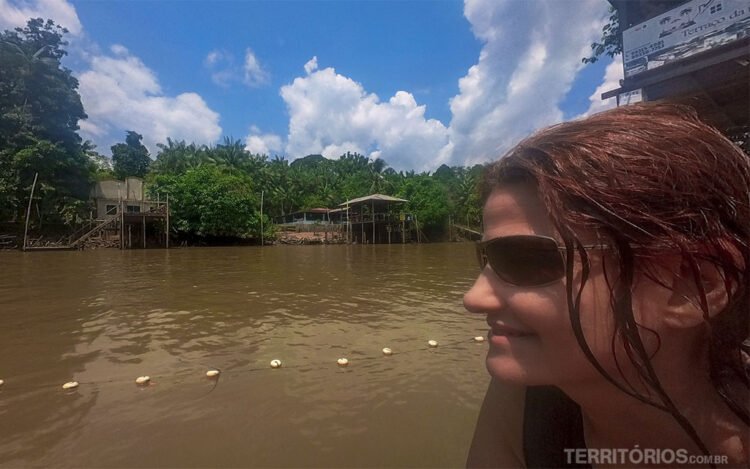 Mulher morena com óculos escuros e cabelo molhado observa o rio em frente. Do outro lado, casas de palafitas em meio a vegetação. Céu azul com nuvens