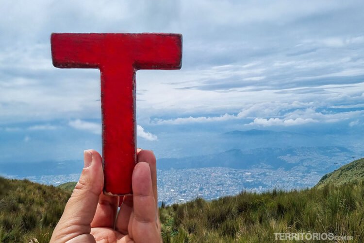 Mão branca segura a letra T vermelha de madeira (Tesão) no alto de uma montanha com vista para a cidade. Céu nublado.