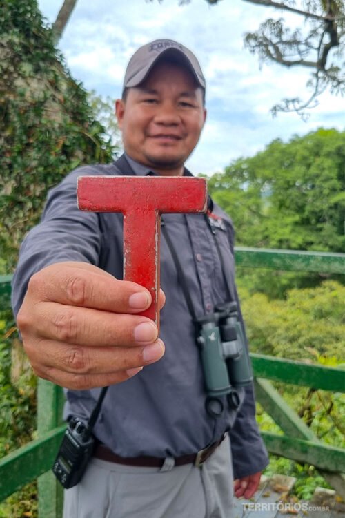 Homem indígena veste uniforme e boné de cor cinza e exibe a letra de vermelha na mão direita. Ele carrega um binóculo no pescoço e rádio na cintura. Ao fundo densa floresta verde e céu nublado. 