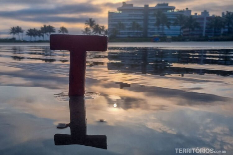 Letra T vermelha de madeira na beira da praia com reflexo do céu na água do mar, ao fundo prédios, palmeiras e céu nublado.