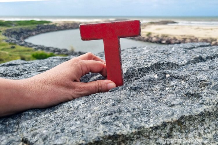 Letra T vermelha de madeira sobre rocha, ao fundo riacho deságua no mar e o céu está nublado.