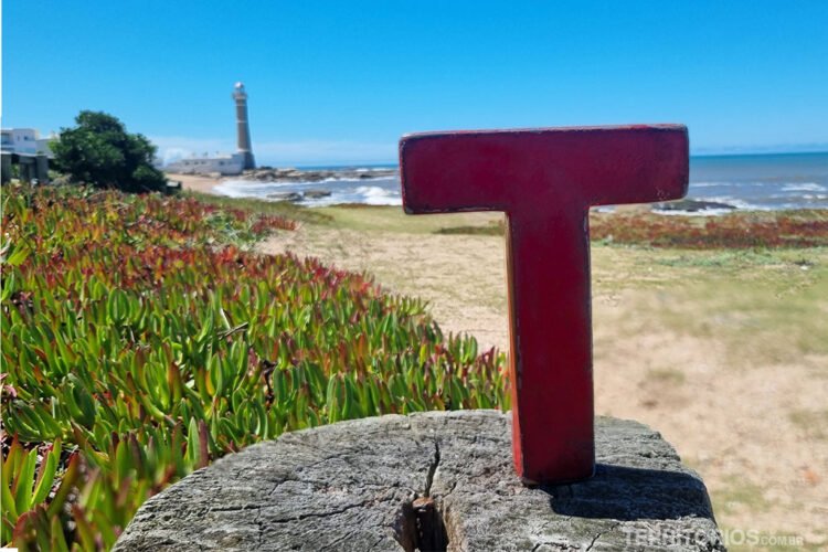 Letra T vermelha (Tesão) sobre tronco de madeira na praia, ao fundo vegetação, areia, céu azul e farol.