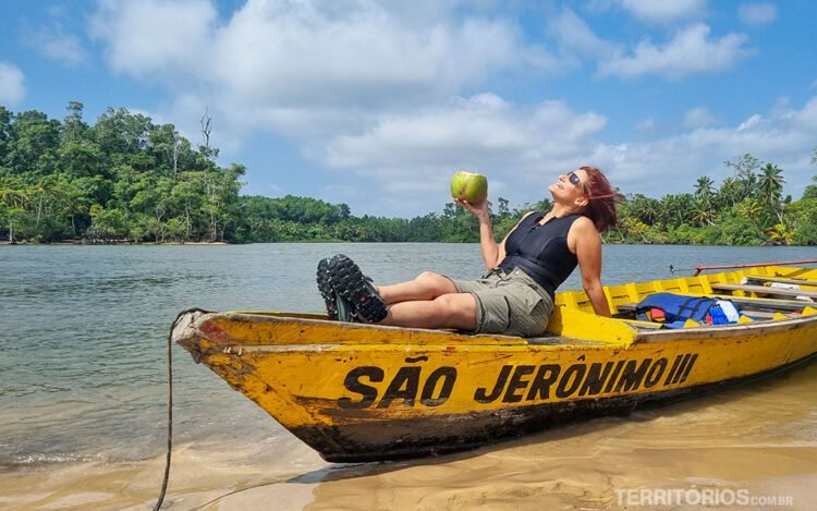 Mulher de óculos escuro sorri olhando para cima vestida com body preto, bermuda bege e tênis sentada em barco na praia de um rio da Ilha de Marajó. Ao fundo mata verde e céu azul com nuvens.