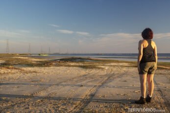 Mulher morena veste bermuda marrom e blusa preta. Observa o fim de tarde na Lagoa Mirim