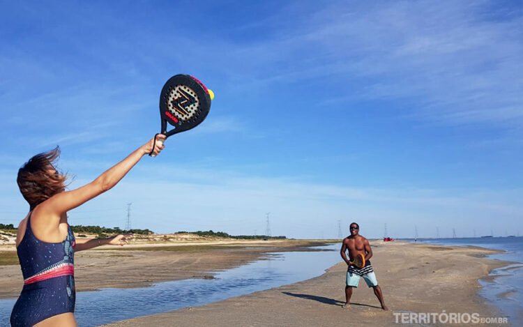 Mulher branca de maiô rebate bola com raquete em cenário de praia tranquila. Homem negro aguarda ao fundo. Céu azul com nuvens.