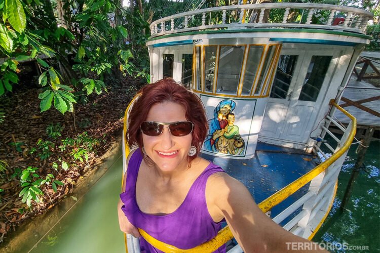 Mulher de óculos escuros e blusa roxa sorri na proa de barco típico da Amazônia.
