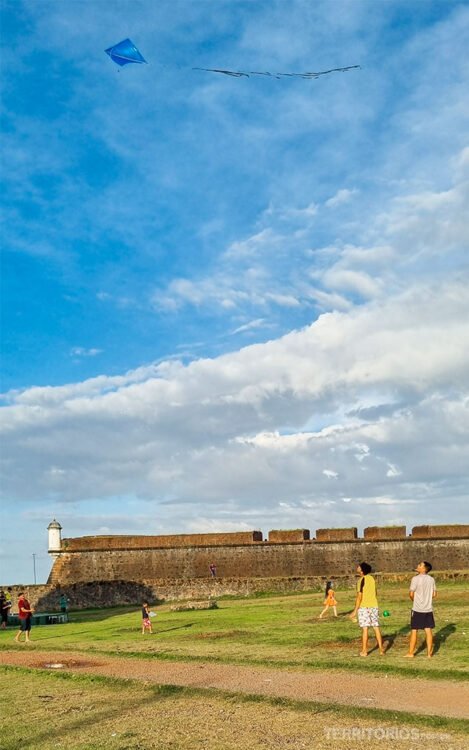 Crianças brinca com pipa azul no céu com nuvens e em frente a muralha da fortaleza de pedra.