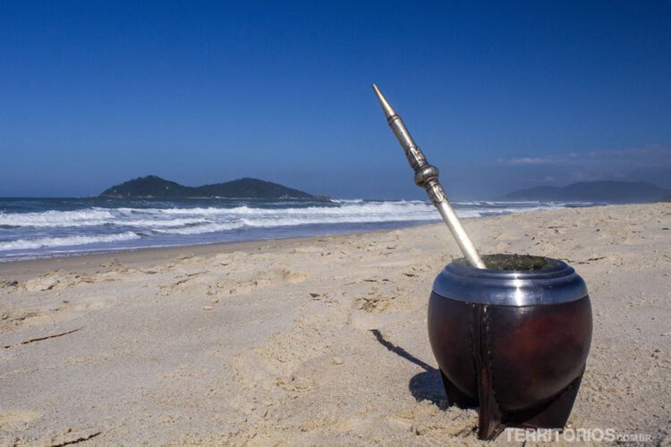 Cuia de chimarrão servido na areia da praia do Rio Tavares em frente a Ilha do Campeche, céu azul.