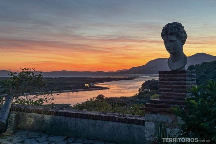 Estátua de cabeça grega em pedestal, um muro e vista para rio, montanhas, mar e céu na hora do crespúsculo.